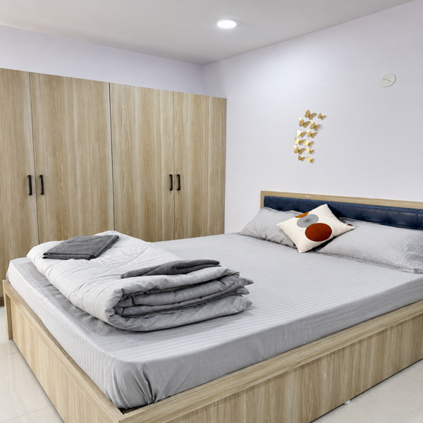 Modern bedroom, contemporary bedroom, cozy bedroom, master bedroom, king size bed, wooden bed, light wood furniture, wooden wardrobe, built-in wardrobe, spacious wardrobe, bedroom storage, upholstered headboard, blue headboard, grey bedsheet, white bedding, folded blanket, grey blanket, decorative pillows, accent pillow, minimalist decor, modern interior, elegant bedroom, clean bedroom, bright bedroom, neutral colors, beige wood, white walls, recessed ceiling light, ceiling lighting, wall decoration, butterfly wall decor, marble flooring, tiled floor, bedroom furniture, home decor, interior design, residential interior, modern home, stylish bedroom, comfortable bed, organized bedroom, soft bedding, contemporary furniture, storage cabinet, luxury home, peaceful bedroom, simple decor, Indian home interior, apartment bedroom, real estate interior, bedroom photography, warm wood tones.